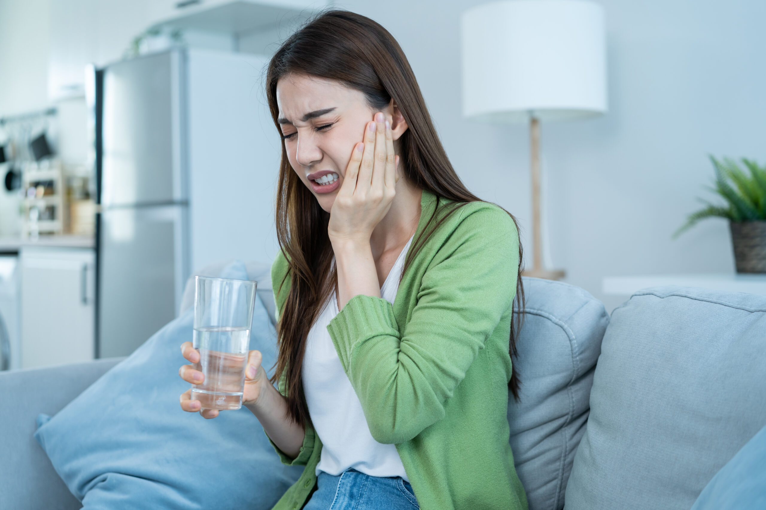 Asian beautiful woman feel terrible toothache after drink cold water. Attractive female sit on sofa in living room touching cheek, feel hurt and suffering from sensitive tooth ache, pain and cavities.