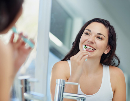 Smiling woman brushing her teeth in front of a mirror as part of her daily routine to support dental cleaning in Cary NC